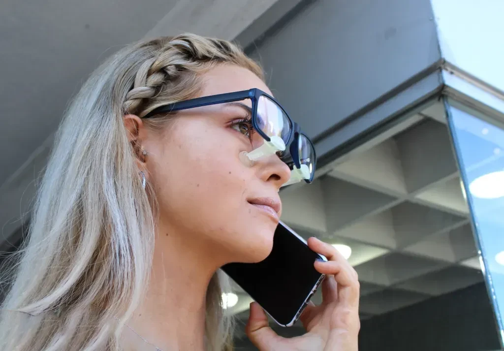 تصویری از خانمی در حال حرف زدن با گوشی و استفاده از نگهدارنده عینک بعد از عمل بینی